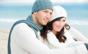 Young couple wearing beanies sit at the beach looking close and happy